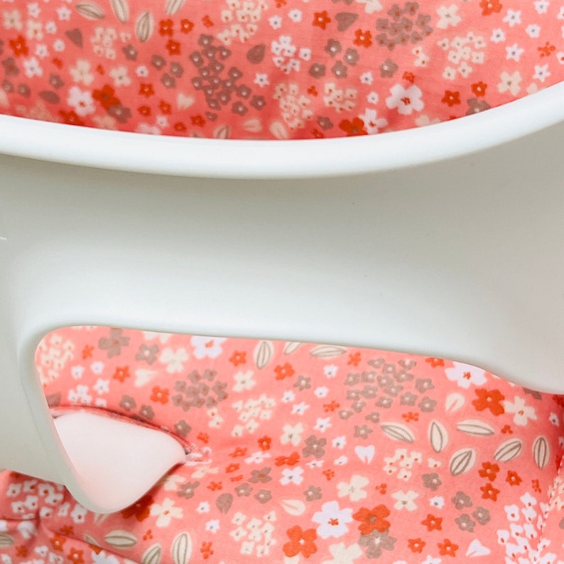 May include: A close-up of a white high chair with a pink floral fabric seat cover. The fabric has a small-scale floral pattern with pink, white, and brown flowers.