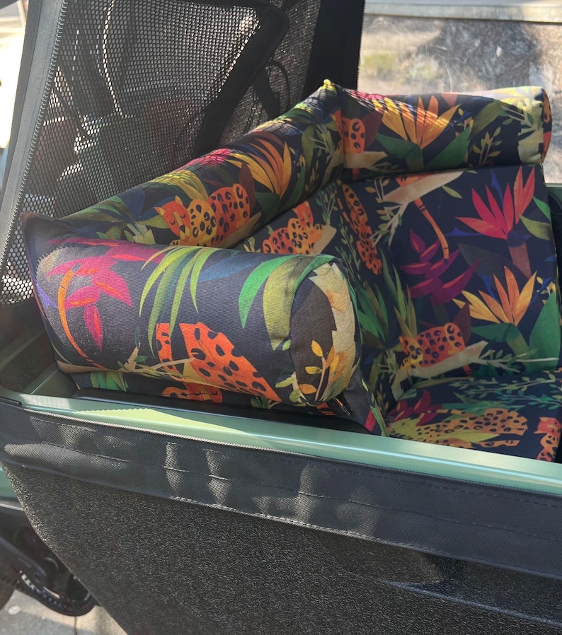 May include: A cargo bike seat with a dark background and a vibrant tropical floral pattern. The seat features a combination of orange, yellow, red, and green leaves and flowers. The seat is designed for comfort and style.