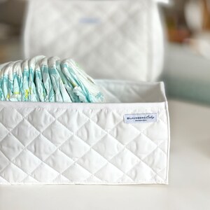 May include: A white quilted storage bin with a label that reads "Blausberg Baby Hamburg" and is filled with a stack of baby diapers.