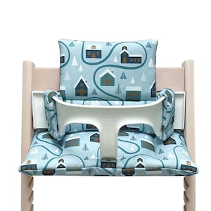 May include: A blue and white high chair cushion with a winter village pattern. The cushion features a white strap and a white back support.