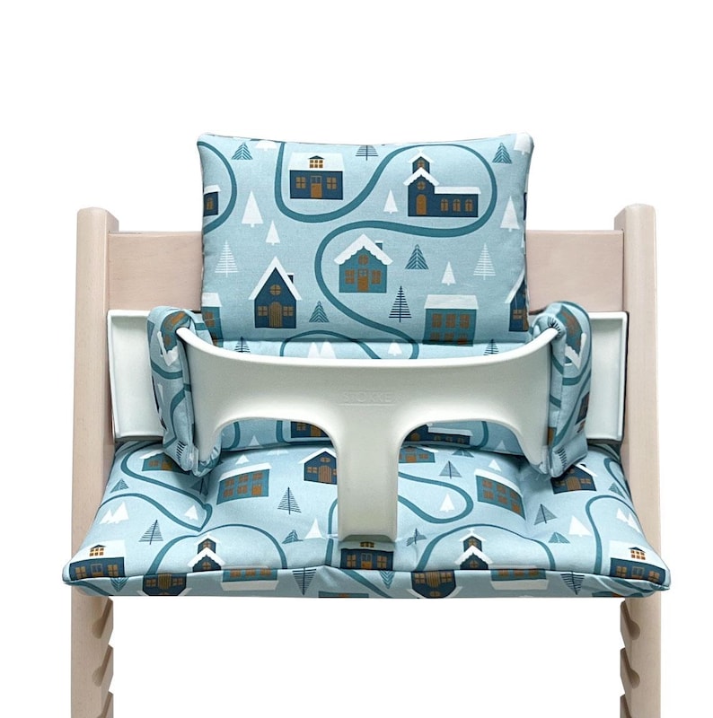 May include: A blue and white high chair cushion with a winter village pattern. The cushion features a white strap and a white back support.