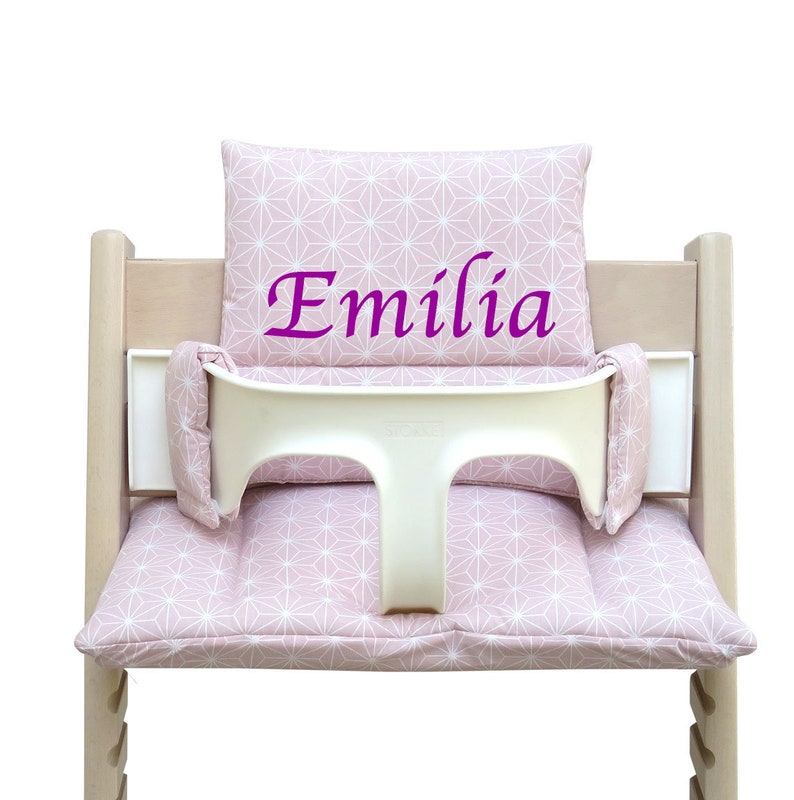 Pu&ograve; includere: Un cuscino per seggiolone rosa e bianco con motivo a stelle e un cuscino schienale abbinato. Il cuscino ha il nome "Emilia" ricamato in viola.