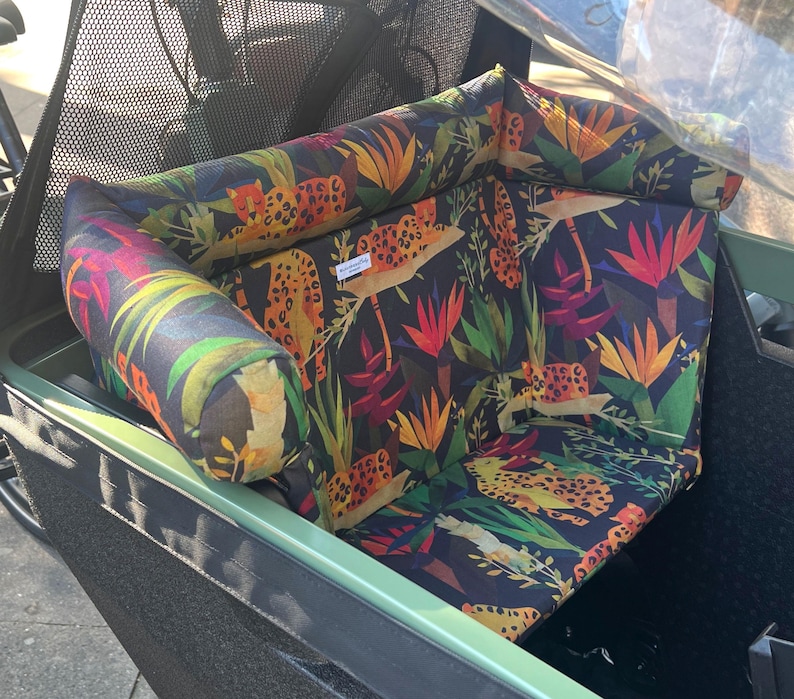 May include: A patterned seat cushion and bolster pillow set featuring a jungle theme with leopards, tropical flowers, and foliage in shades of orange, green, and red against a dark background. The set is designed to fit a cargo bike.
