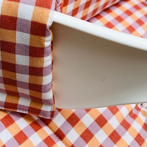 May include: Close-up of a red and orange gingham fabric with white squares. The fabric is folded and has a white seam around the edges.