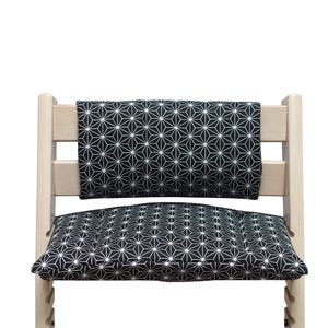 K&ouml;nnte beinhalten: Ein helles Holzhochstuhl mit einem schwarz-wei&szlig;en geometrischen Muster auf Kissen und R&uuml;ckenlehne. Das Muster zeigt wei&szlig;e sternf&ouml;rmige Formen auf schwarzem Hintergrund. Das Design des Stuhls ist modern und minimalistisch.