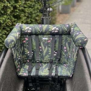 Ein Cargobike-Sitz mit floralem Muster. Das Kissen ist schwarz mit einem Muster aus rosa Blumen und grünen Blättern. Er hat schwarze Gurte und ist für ein Kind konzipiert. Das Kissen verfügt über ein gutes, dickes und schützendes Nackenpolster, an das sich Kinder anlehnen können, falls sie einmal bei der Fahr einschlafen.