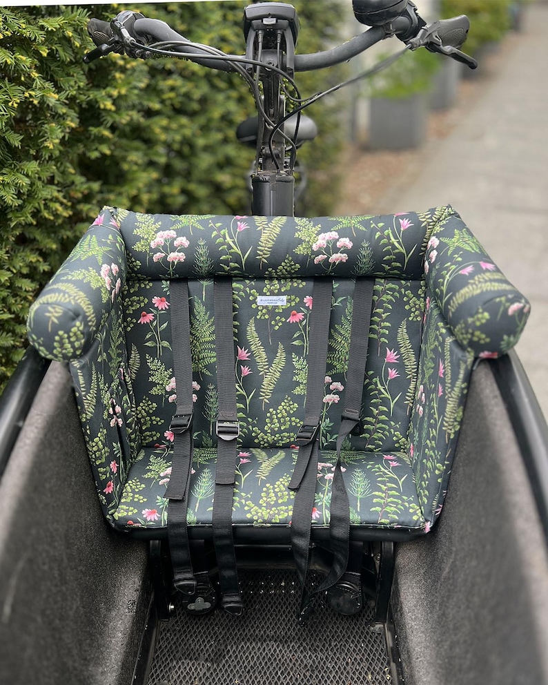 Ein Cargobike-Sitz mit floralem Muster. Das Kissen ist schwarz mit einem Muster aus rosa Blumen und grünen Blättern. Er hat schwarze Gurte und ist für ein Kind konzipiert. Das Kissen verfügt über ein gutes, dickes und schützendes Nackenpolster, an das sich Kinder anlehnen können, falls sie einmal bei der Fahr einschlafen.
