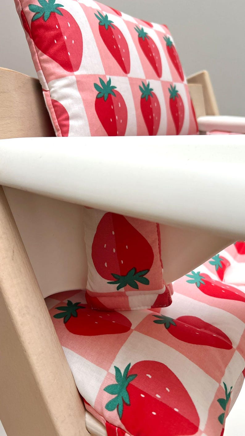 May include: A white high chair with a pink and white gingham pattern cushion and a matching pillow featuring a strawberry print.