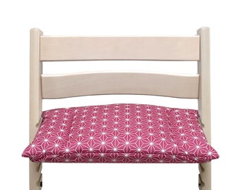 May include: A light-colored wooden high chair with a magenta seat cushion. The cushion features a white geometric starburst pattern. The chair is designed for a child and is set against a white background.