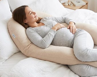 May include: A long, U-shaped body pillow in a solid light beige color with a textured muslin fabric cover. The pillow curves around the torso and legs, providing support along the side of the body, and features a small white brand label on the side.