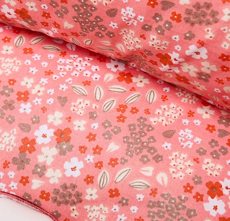 May include: Pink fabric with a floral pattern featuring small white and orange flowers, leaves, and a few hearts. The fabric has a textured surface.