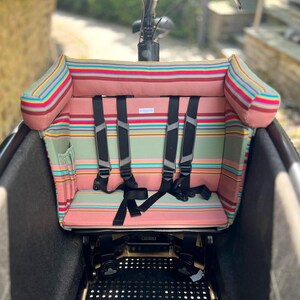 Sitzkissen für das Urban Arrow Family oder Lovens Lastenrad. Man sieht ein gestreiften Stoff mit gepolsterter Kopfstütze und einem Fünf-Punkt-Gurtsystem. Das Kissen für das Cargobike is rosa salbeifarben gestreift