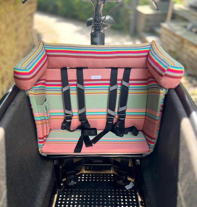Sitzkissen für das Urban Arrow Family oder Lovens Lastenrad. Man sieht ein gestreiften Stoff mit gepolsterter Kopfstütze und einem Fünf-Punkt-Gurtsystem. Das Kissen für das Cargobike is rosa salbeifarben gestreift