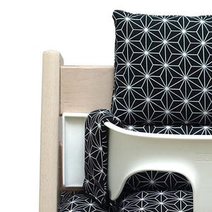 May include: A white high chair with a black and white geometric patterned cushion and back support. The chair has a wooden frame and a white tray. The cushion and back support feature a repeating starburst design.