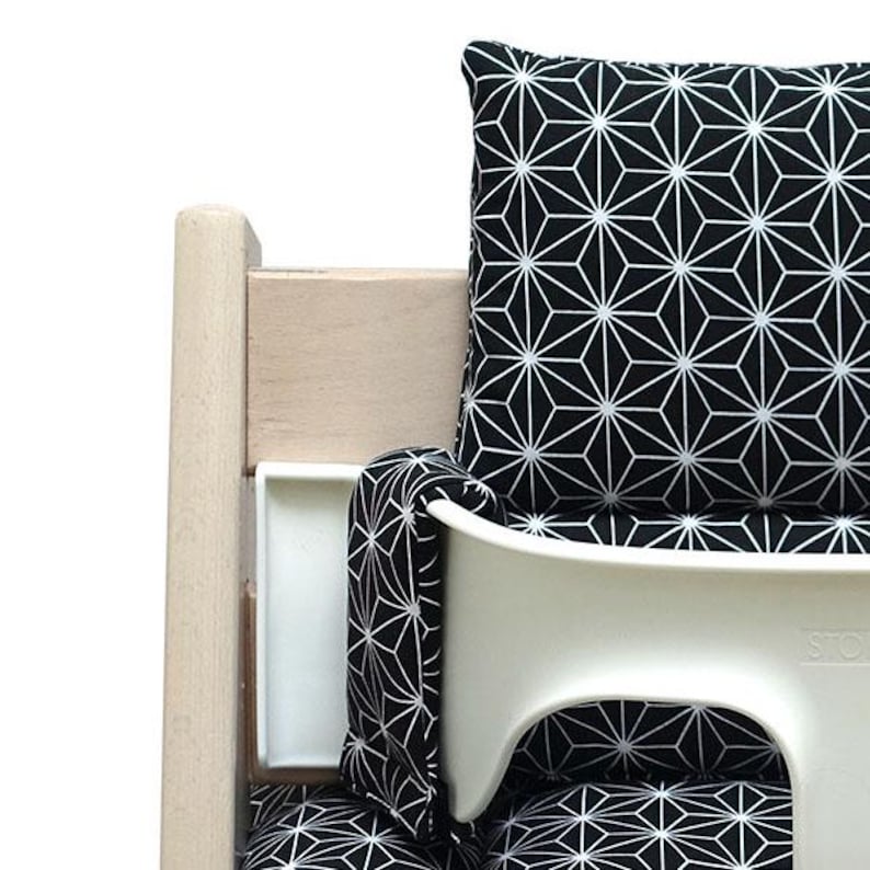 May include: A white high chair with a black and white geometric patterned cushion and back support. The chair has a wooden frame and a white tray. The cushion and back support feature a repeating starburst design.