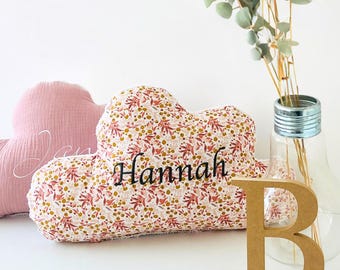 May include: A pink and white cloud-shaped cushion with the name "Hannah" embroidered on it. The cushion is decorated with a floral pattern in shades of pink, white, and gold.