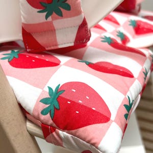 May include: A pink and white checkered high chair cushion with a strawberry pattern. The cushion is made of soft fabric and has a comfortable design.
