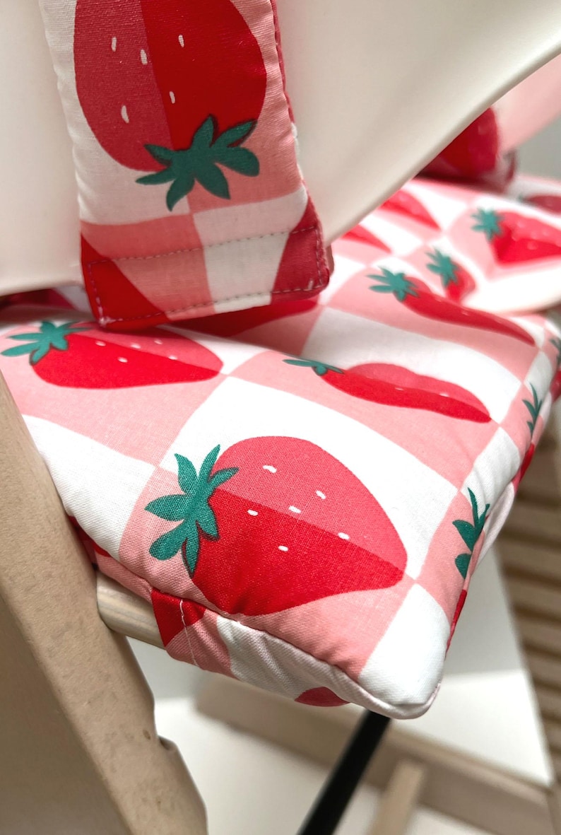 May include: A pink and white checkered high chair cushion with a strawberry pattern. The cushion is made of soft fabric and has a comfortable design.