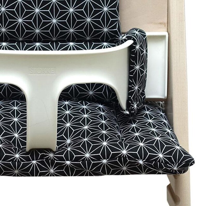 May include: A white high chair with a black and white geometric patterned cushion and backrest. The high chair has a white tray and the brand name "STOKKE" is visible on the chair.
