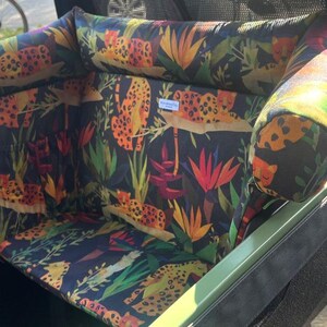 May include: A cargo bike seat cushion set with a jungle-themed pattern. The fabric features orange leopards, green leaves, and colorful flowers on a dark blue background. The set includes a seat and a headrest.