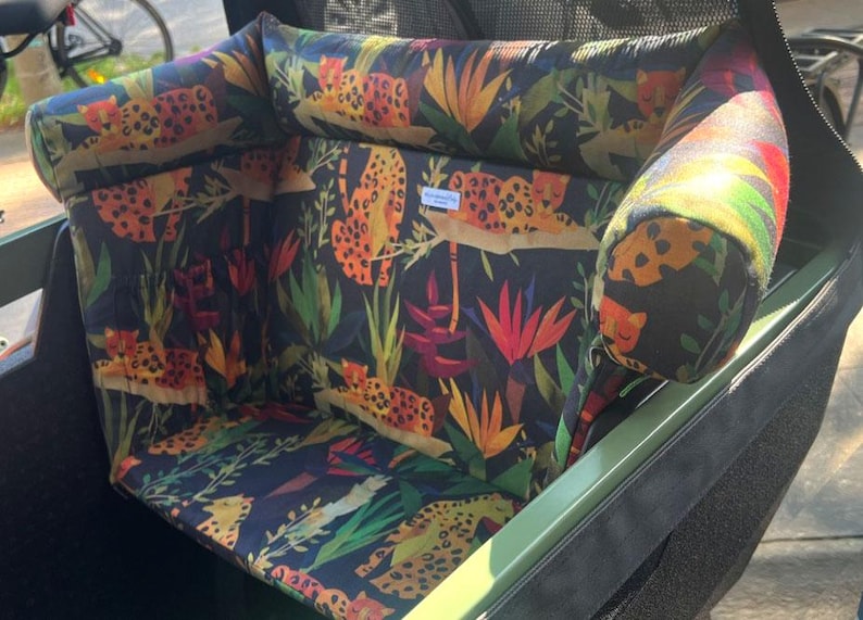 May include: A cargo bike seat cushion set with a jungle-themed pattern. The fabric features orange leopards, green leaves, and colorful flowers on a dark blue background. The set includes a seat and a headrest.