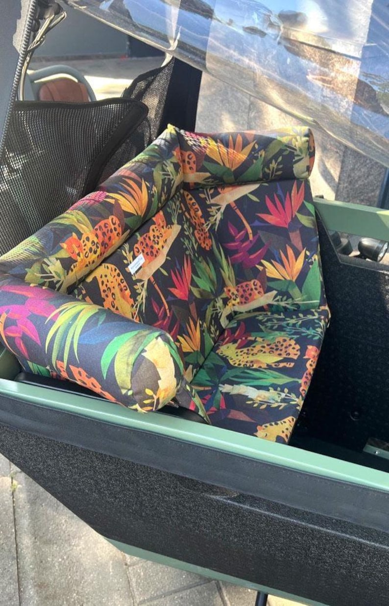 May include: A stroller seat and bolster pillows feature a vibrant jungle print with leopard spots, tropical flowers, and lush green foliage on a dark background. The seat and pillows are inside a green stroller.