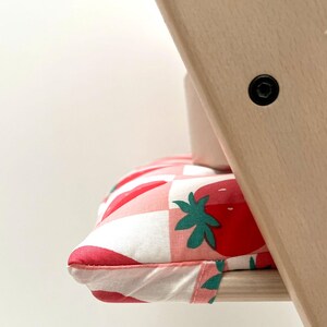 May include: A pink and white checkered chair cushion with a strawberry pattern. The cushion is made of soft fabric and has a white border.