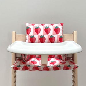 May include: A white high chair with a pink and red strawberry patterned cushion and a white tray.