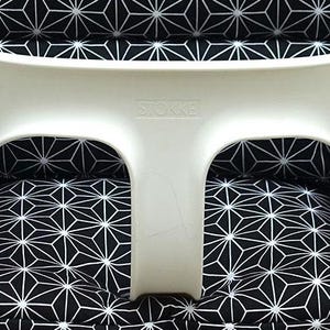 May include: A white high chair with a black and white geometric patterned cushion. The high chair has the brand name "Stokke" printed on the front. The cushion features a repeating starburst design.