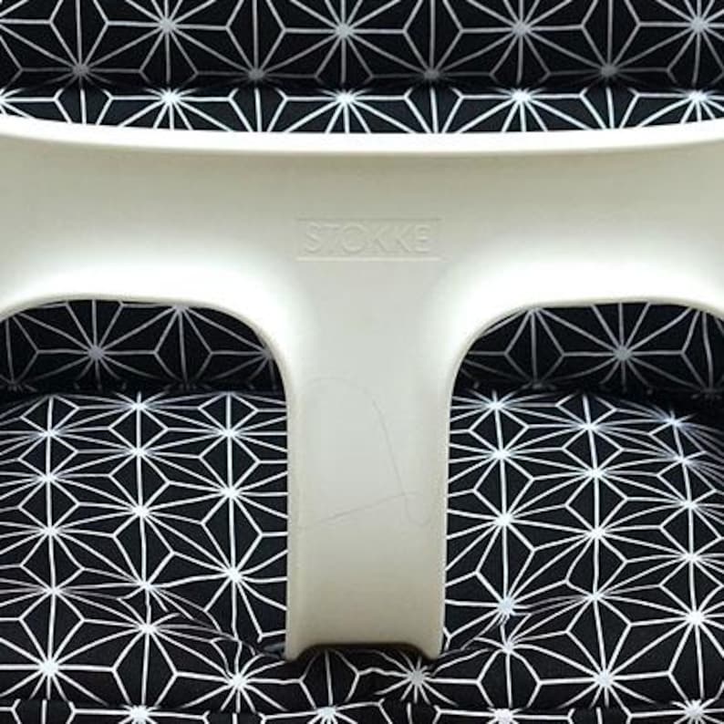 May include: A white high chair with a black and white geometric patterned cushion. The high chair has the brand name "Stokke" printed on the front. The cushion features a repeating starburst design.