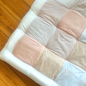 Könnte beinhalten: Eine quadratische, gesteppte Matte mit weißem Rand. Die Mitte ist ein Patchwork aus verschiedenen Stoffen in Rosa-, Creme- und Weißtönen. Einige Quadrate haben florale Muster, andere sind uni oder strukturiert.