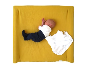 May include: A baby wearing a white shirt and dark blue pants is lying on a yellow changing pad. A white muslin swaddle blanket is on the pad.