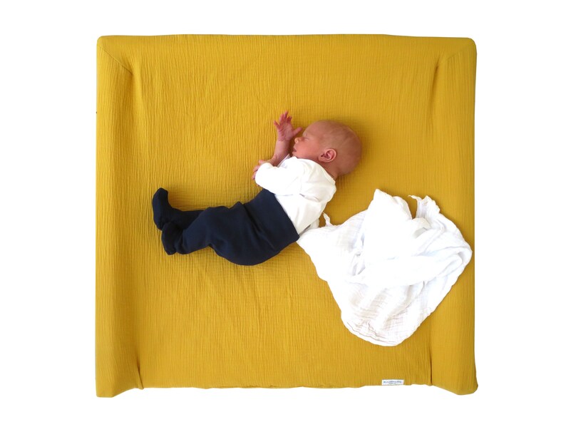 May include: A baby wearing a white shirt and dark blue pants is lying on a yellow changing pad. A white muslin swaddle blanket is on the pad.