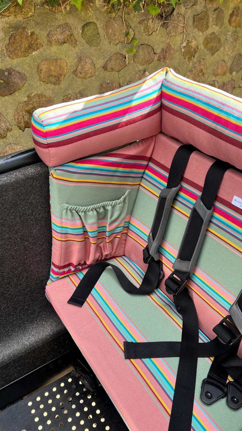 Das rosa-grün-salbei gestreifte Kissen für das Urban Arrow Lastenrad. Das  Lastenrad Kissen für das Urban Arrow wird von der Seite gezeigt. Es verfügt über eine Tasche, in die Kinder während der Fahrt im Lastenrad z.B. eine Trinkflasche stecken können. Es hat eine Nackenrolle, die gut gepolstert und angenehm ist und an die Kinder ihren Kopf sicher lehnen können, wenn sie während der Fahrt im Cargo Bike einschlafen.