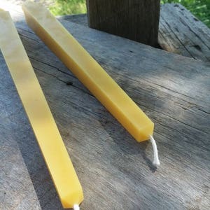 May include: Two long, yellow beeswax candles with white wicks. The candles are lying on a wooden surface.