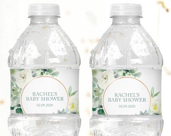May include: Two clear plastic water bottles with white labels. The labels feature a watercolor design of white flowers and green leaves with a gold circle around the text "Rachel's Baby Shower 02.09.2020".