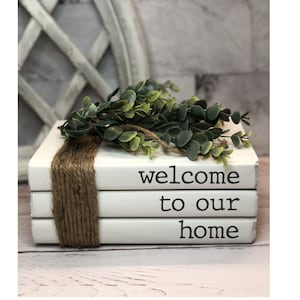 May include: A stack of three white books with the words "welcome to our home" written on them. The books are decorated with a sprig of green foliage and twine.