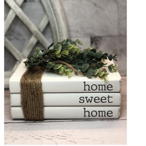 May include: A stack of three white painted books tied with twine, with the words "home sweet home" printed on the sides. A green leafy garland rests on top, creating a rustic, farmhouse aesthetic.