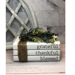 May include: A stack of three white books with the words "grateful", "thankful", and "blessed" printed on them. The books are tied together with twine and decorated with a sprig of green foliage.