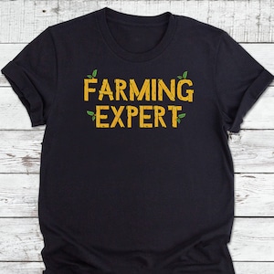 以下が含まれることがあります： 緑の葉のアクセントが付いた黄色の「FARMING EXPERT」という文字が書かれた黒いTシャツです。