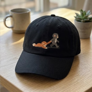 Può includere: Un cappellino da baseball nero con la grafica di una scimmia grigia che cammina trascinando per il braccio una scimmia arancione più piccola sul terreno. Il cappellino presenta una visiera curva e una corona strutturata con occhielli cuciti.