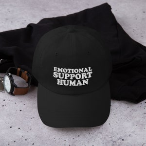 May include: A black baseball cap with white embroidered text that reads "Emotional Support Human".