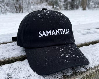 Könnte beinhalten: Eine schwarze Baseballkappe mit dem weiß gestickten Text SAMANTHA? auf der Vorderseite. Die Kappe hat einen gebogenen Schirm und eine strukturierte Krone, wobei kleine Belüftungsösen auf den oberen Stoffbahnen zu sehen sind.