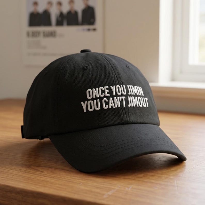 K&ouml;nnte beinhalten: Eine schwarze Baseballkappe mit wei&szlig;em, gesticktem Text auf der Vorderseite, der ONCE YOU JIMIN YOU CAN'T JIMOUT lautet. Die Kappe hat eine strukturierte Krone, einen gebogenen Schirm und einen Knopf oben in der Mitte f&uuml;r ein schlichtes Design.