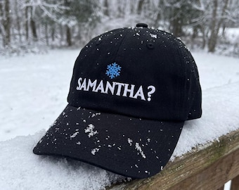 Könnte beinhalten: Eine schwarze Baseballkappe mit einem gestickten hellblauen Schneeflockensymbol über dem weißen Schriftzug SAMANTHA?, der mittig auf der Vorderseite platziert ist. Die Kappe ist mit kleinen Schneeflocken bedeckt, was auf eine Winterumgebung hindeutet.