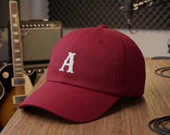 Könnte beinhalten: Eine kastanienbraune Baseballkappe mit einem weißen, gestickten Buchstaben A auf der Vorderseite. Die Kappe hat einen gebogenen Schirm, eine strukturierte Krone mit Belüftungsösen und einen Stoffriemenverschluss auf der Rückseite.