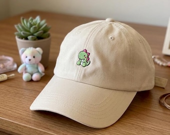 May include: A beige baseball cap featuring a small, embroidered green dinosaur with pink spikes on the front panel. The cap has a structured crown, a curved brim with visible stitching, and a button at the top center of the fabric dome.