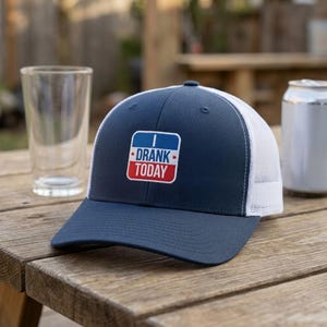 May include: A navy blue baseball cap with a white mesh back and a rectangular patch on the front. The patch features a blue top section with the white text I, a white middle section with the blue text DRANK, and a red bottom section with the white text TODAY.