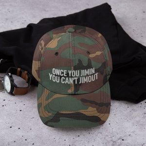 K&ouml;nnte beinhalten: Eine Baseballkappe mit Camouflage-Muster und dem aufgestickten wei&szlig;en Text "ONCE YOU JIMIN YOU CAN'T JIMOUT" auf der Vorderseite. Die Kappe hat eine strukturierte Krone, einen gebogenen Schirm und einen Knopf oben in klassischen Gr&uuml;n-, Braun- und Schwarzt&ouml;nen.