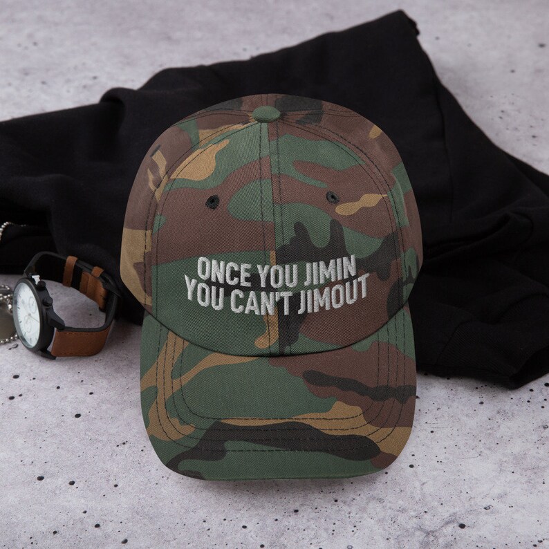 K&ouml;nnte beinhalten: Eine Baseballkappe mit Camouflage-Muster und dem aufgestickten wei&szlig;en Text "ONCE YOU JIMIN YOU CAN'T JIMOUT" auf der Vorderseite. Die Kappe hat eine strukturierte Krone, einen gebogenen Schirm und einen Knopf oben in klassischen Gr&uuml;n-, Braun- und Schwarzt&ouml;nen.
